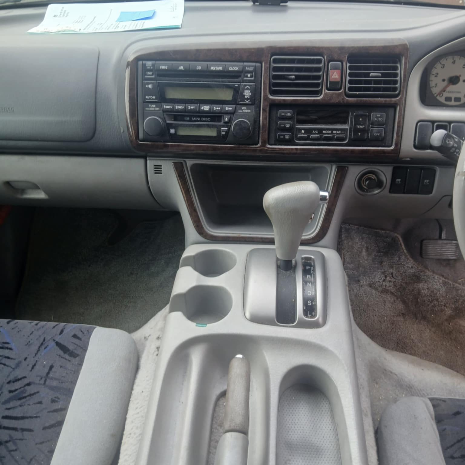Interior view of a Mazda Bongo, showing the dashboard, centre console, and gear selector. The panel features a built-in radio and CD player with classic buttons and dials, air conditioning controls, cup holders, and storage compartments, alongside an automatic gear shifter and handbrake